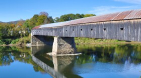 The Cornish-Windsor covered bridge spans the Connecticut River on a bright blue day, with foliage in the background