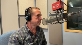 A man in a button down plaid shirt wears headphones and sits in front of a radio microphone