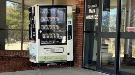 A vending machine next to the entrance of an office building. The machine has a glass front, behind which boxes of the overdose-reversal medication naloxone are visible. The exterior of the vending machine is white with different messages on it, including "Save one life."