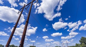 A tall power line towers in the foreground, in front of a blue sky dotted with clouds. 