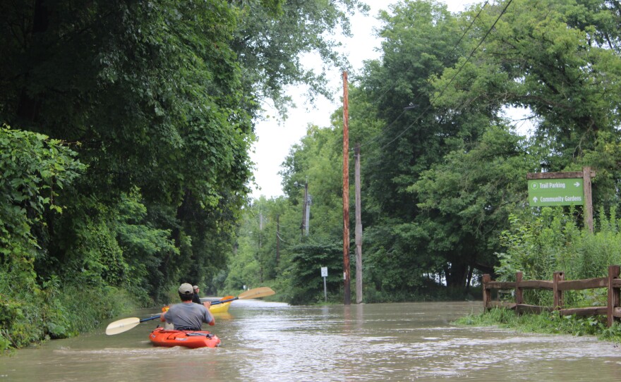 Two kayakers, facing away from the camera, paddle on a flooded road.