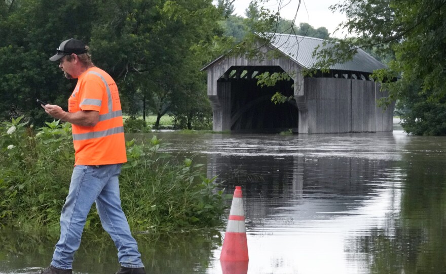 A man wearing a bright orange safety shirt walks while holding a phone next to floodwaters. In the background is a covered bridge