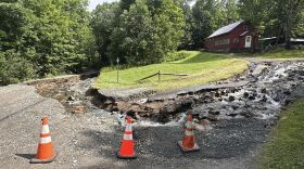 Three orange traffic cones are set up in front of a rode that's eroded down the middle with water flowing down it.