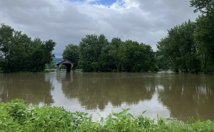 Floodwaters surround one end of a covered bridge, seen in the distance amid trees.