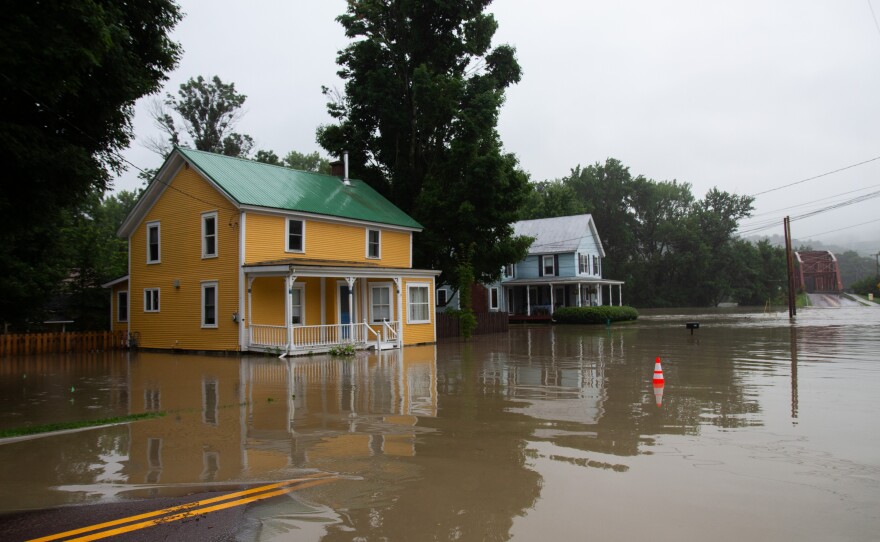 Brown water covers a paved road and surrounds a bright yellow house. An orange safety cone sits in the middle of the water.