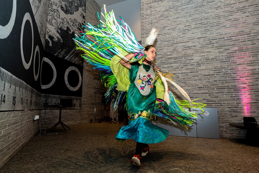 A photo of a person inside a grey brick room. The person is wearing a green and blue dress with a lime green shawl with many long tassels in all different shades of green and blue. The person is mid-dance, so the shawl tassels are spread out like bird wings. 