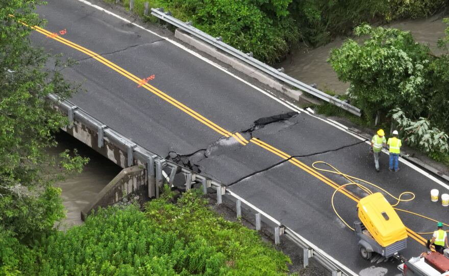 Two people in yellow construction safety equipment stand near a crack in a bridge over water. A construction vehicle is parked nearby.