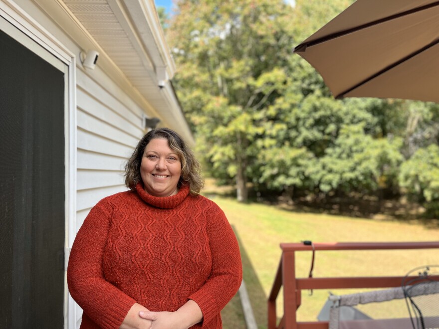 A woman stands on a deck outside her home, with grass and trees in the background.