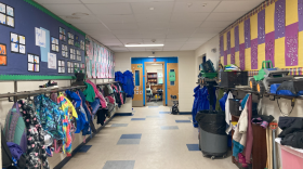 Bags, jackets and raincoats hang on both sides of the hallway. Colorful art made by the kindergarten classes is plastered on the walls. At the end of the hall is a classroom with open doors. 