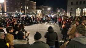 A large group of people stand in a circle on a plaza in downtown Concord, some holding candles