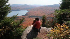 A parent and child on a rocky ledge overlooking a lake and mountains covered in bright orange fall foliage.