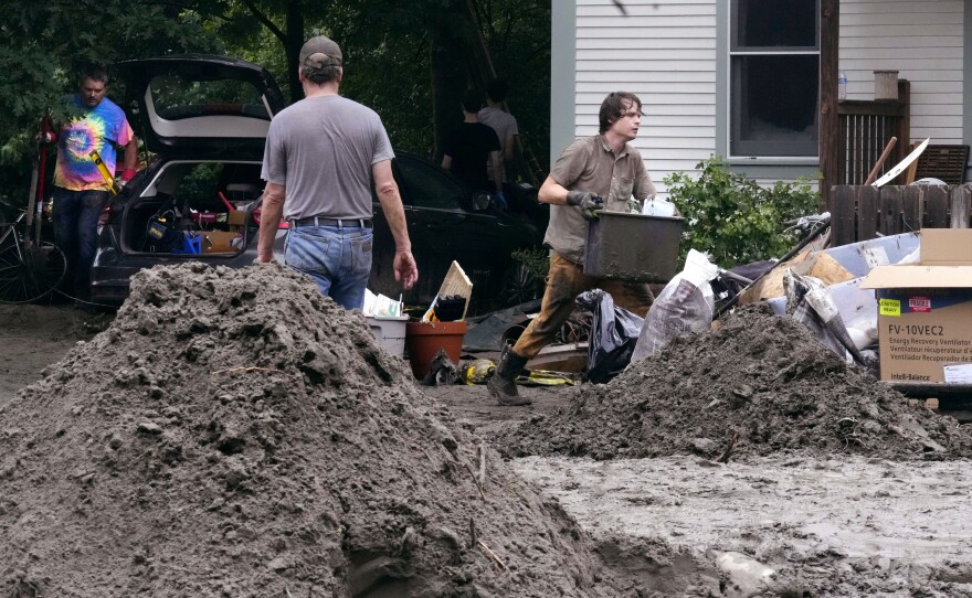 A man carries a bin of items outside a home, surrounded by piles of dirt and mud and belongings