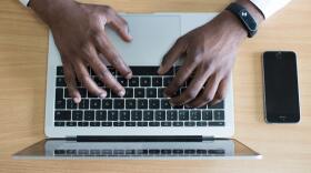 A top-down photo of hands on a computer keyboard. An iPhone sits to the side on a wooden table. 