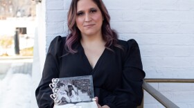 A woman in a black dress sits against a white brick wall with a somber expression on her face. She holds a black and white photo of a couple.