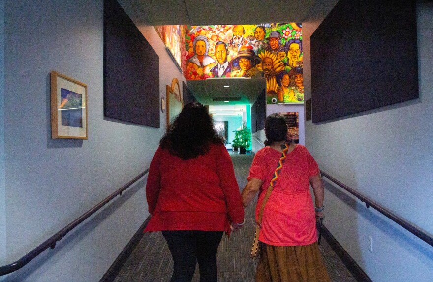 A photo of two women seen from the back, one in a red shirt with long black hair, and one in a pink shirt with a grey braid over her shoulder. They're holding hands inside a building hallway, and a mural of people of color is visible in the distance.