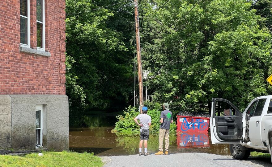 Two people stand on a pavement road between a red brick building and a white truck with the driver side door open. They stand facing dark colored water that's approaching the building through a tree-covered area.