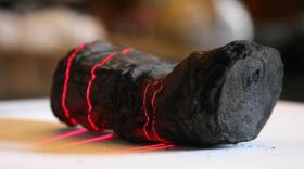 A charred, lumpy papyrus scroll, with red lasers giving the sense of the scroll being scanned by an X-Ray. This scroll was recovered from a Roman villa, carbonized by scorching volcanic gas almost two thousand years ago. 