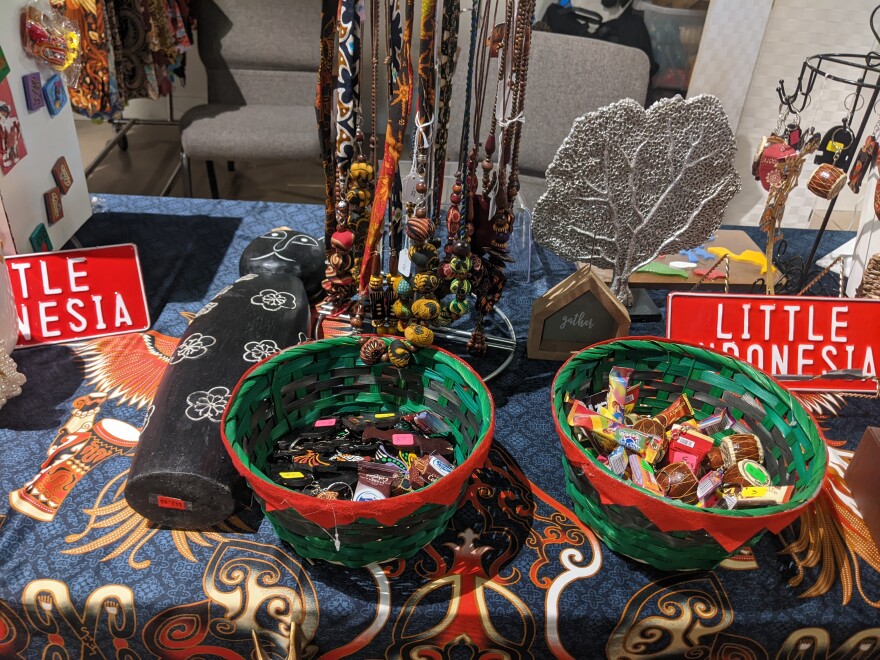 A close up of a vendor booth that has two treat baskets, a cat ornament, beads, a leaf statue and a sign that says "Little Indonesia".jpg