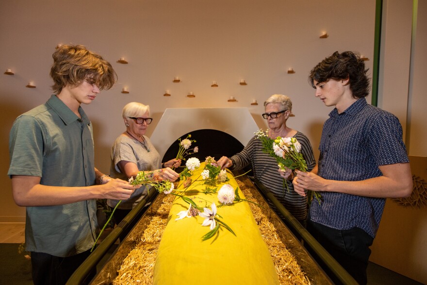 Guests demonstrate placing flowers on a shrouded mannequin in Recompose's Gathering Space. Decedents here are surrounded by wood chips and alfalfa, and placed in a special vessel that encourages decomposition.