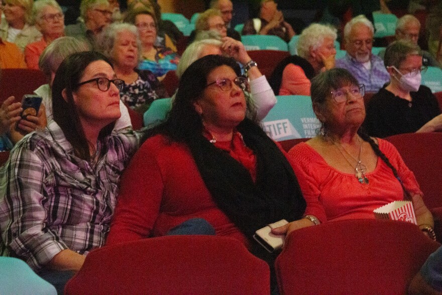 A photo of three women, two younger, one older, all in glasses, sit in a movie theater surrounded by people. They're all looking up, as if toward a screen.