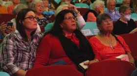 A photo of three women, two younger, one older, all in glasses, sit in a movie theater surrounded by people. They're all looking up, as if toward a screen.