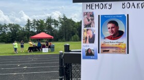  In the foreground is a white poster titled Memory Board. An image of a young man is in the center and three smaller images of other people are along the sides. The larger image reads "Forever in our Hearts: David Robert Plaisted, July 15, 1992 - January 31, 2022." Behind the poster is an outdoor track. Several people are gathered around a red tent in the grass in the center of the track.  