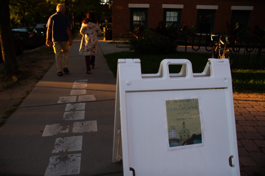 A photo of a white sandwich board with a smaller poster reading "Sugarcane" over the image of a church. Behind the sandwich board are two people on a sidewalk, where deep golden sunlight is shining.