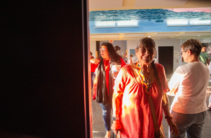 A photo of a woman with glasses and a long braid smiling in a sun-drenched room with people in the background. 