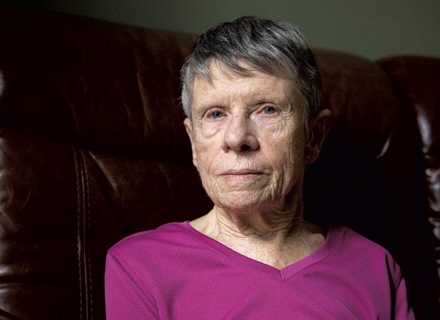 An older white woman with short gray hair and a magenta shirt looks directly at the camera, with a brown leather seat back in the background.