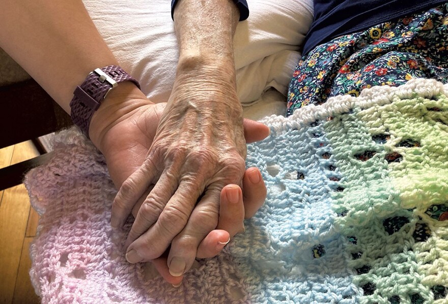 A close-up of the hand of a younger white person holding the hand of an older white person, with a crocheted rainbow blanket in the background.