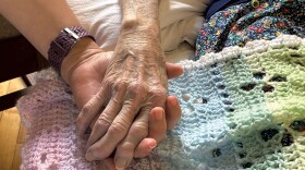 A close-up of the hand of a younger white person holding the hand of an older white person, with a crocheted rainbow blanket in the background.