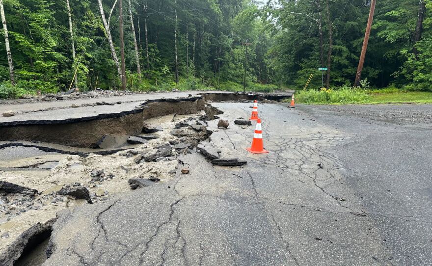 A long crater sits on the left side of a gray pavement road. Orange cones line the missing parts.