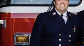 Brian Ryll is a firefighter and the president of the Professional Fire Fighters of New Hampshire. He stands in front of a firetruck in a dress uniform. 