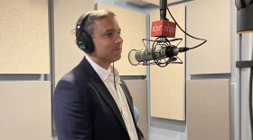 A man in a blue blazer and white button down shirt wears headphones and stands in front of a radio microphone