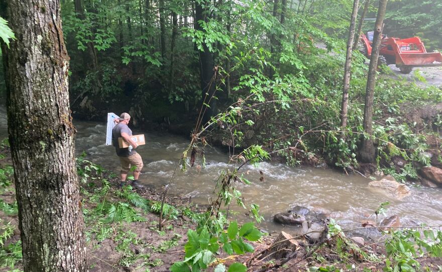 A man wearing a backwards cap and holding items in both hands begins to cross rushing water to the other side, where some pavement and construction vehicles sit.