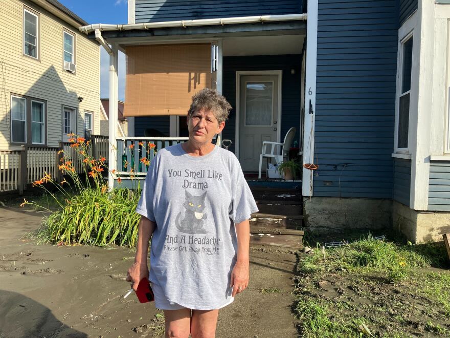 A woman stands in mud outside of a house wearing an oversized T-shirt with a cat on it