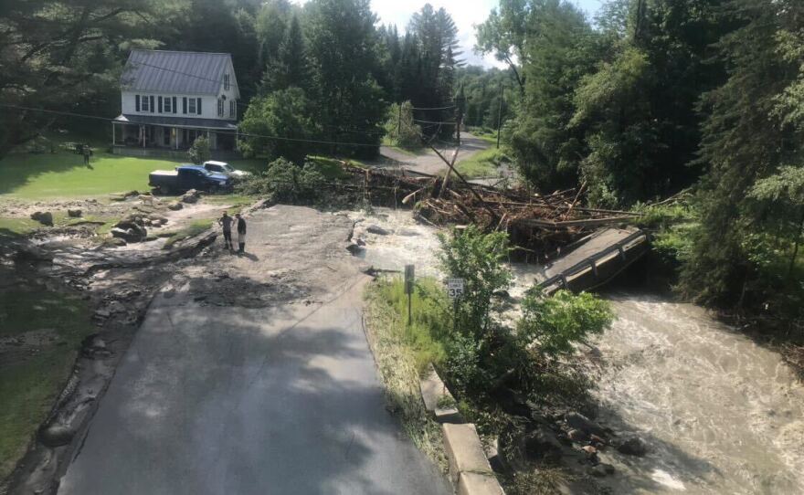 Looking down at a street blocked by downed trees, and parts of it are washed out by murky water flowing perpendicular. Two people stand near by the damage.